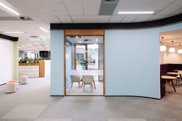 A modern commercial office interior with an open-plan carpeted workspace, pale blue curved partition walls, timber-framed glazed meeting room, and a partially visible breakout or dining area with pendant lighting and booth seating. The space has a suspended acoustic tile ceiling, linear fluorescent lighting, and a bright, professional fit-out.