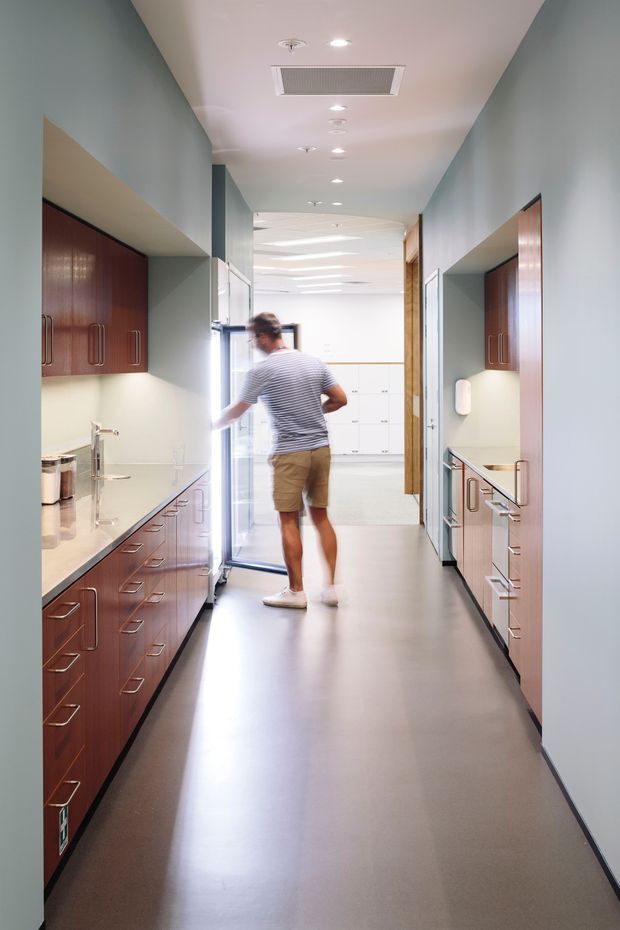 A long, narrow institutional or commercial corridor with built-in timber cabinetry and continuous benchtops on both sides. The cabinetry has dark reddish-brown woodgrain fronts with brushed metal handles, while the walls are muted blue-grey and the floor is a smooth grey resilient finish. Recessed ceiling lights, a large ceiling air-conditioning grille, sinks and work surfaces suggest a staff utility, clinical, or laboratory-related space. A bright glazed doorway opens at the far end, with a person walking through the corridor.