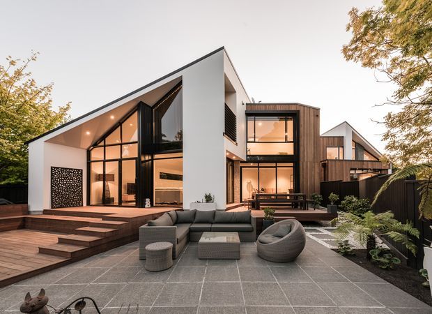 Contemporary multi-level residence with a dramatic pitched roof, extensive floor-to-ceiling glazing, white rendered walls, and a timber-clad upper volume. The home opens onto a broad tiled terrace with outdoor lounge furniture, timber steps, built-in planting, and warm interior lighting visible through the glass.