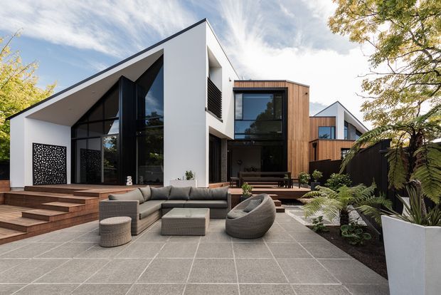 A contemporary multi-level residence with bold geometric rooflines, white rendered walls, dark-framed floor-to-ceiling glazing, and warm timber-clad sections. The landscaped front courtyard features large-format grey paving, tiered timber steps, outdoor lounge furniture, raised planters, and lush ferns.