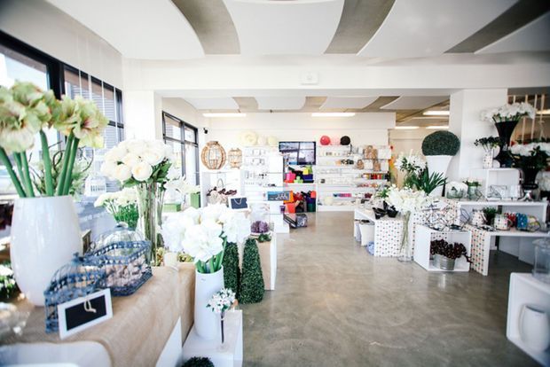 Bright, spacious retail showroom displaying flowers, plants, decorative objects, giftware and homewares. The space has large black-framed windows, a polished concrete floor, white walls, extensive shelving, and distinctive curved ceiling panels with contrasting grey inset sections.