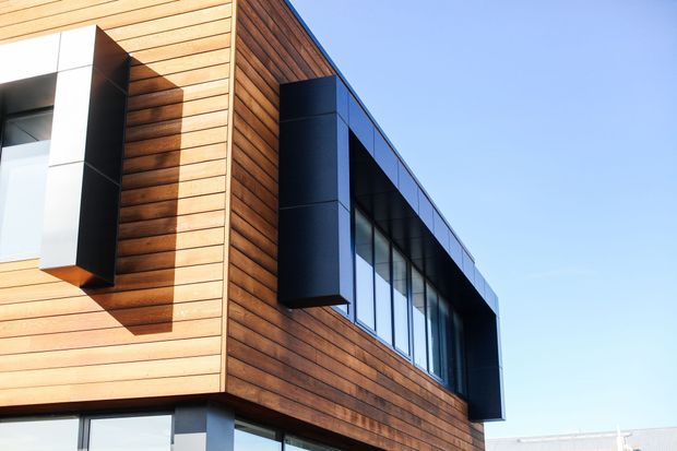 Close-up exterior view of a contemporary multi-storey building with warm horizontal timber cladding, dark projecting window surrounds, and extensive floor-to-ceiling glazing beneath a flat roofline. The composition emphasizes the facade and its crisp geometric detailing against a clear sky.