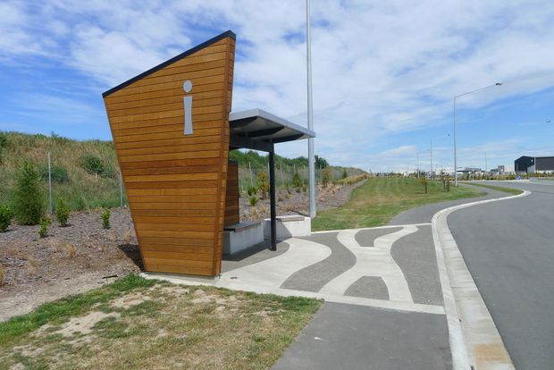Contemporary roadside public information or wayfinding structure clad in horizontal warm timber boards, with a tall angled facade bearing a white information symbol. A small flat-roofed covered shelter and paved patterned pedestrian path sit beside a newly developed roadway and landscaped verge.