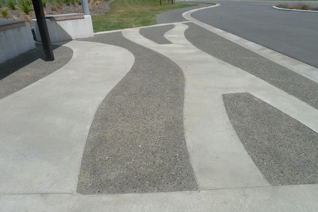 Outdoor paved pedestrian path and driveway edge featuring a flowing pattern of smooth pale concrete bands interspersed with dark exposed-aggregate concrete strips. Low retaining walls, planted areas, and a curving asphalt roadway are visible in the background.