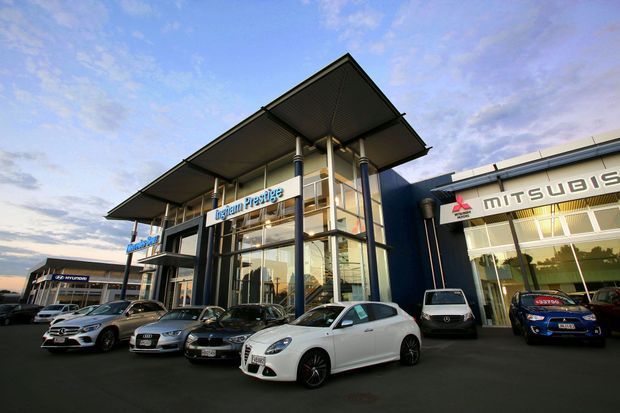Exterior view of a contemporary multi-brand automotive dealership, with a prominent glazed showroom entrance, deep cantilevered canopy roof, dark blue facade sections, and visible Mitsubishi and other automotive branding. Cars are displayed across the forecourt, with adjacent dealership buildings extending along the site.