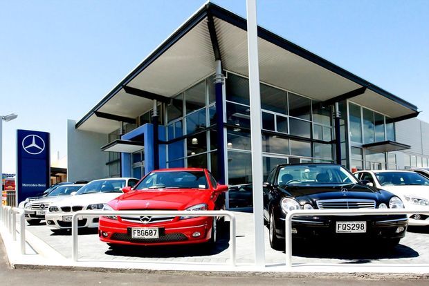 Exterior view of a modern Mercedes-Benz car dealership showroom, with a glazed two-storey commercial facade, projecting flat roof canopy, prominent structural columns, and multiple vehicles displayed in front. The image is brightly lit with a clean, contemporary commercial architectural appearance.