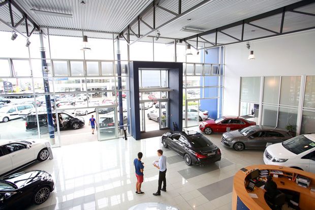 Bright, expansive automotive showroom with full-height glazing, multiple display vehicles, a glossy tiled floor, a curved timber-toned reception desk, and an exposed steel-framed industrial-style ceiling. The showroom opens onto a vehicle forecourt visible through the glazing.