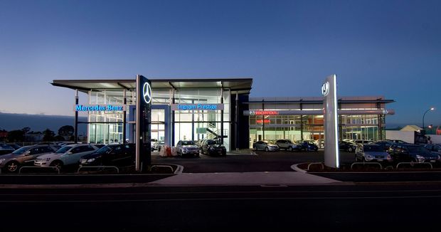 Dusk exterior view of a contemporary Mercedes-Benz automotive dealership, with extensive glass showroom façades, illuminated brand signage, broad flat projecting rooflines, freestanding illuminated sign pylons, and parked vehicles in front. The commercial building is the primary subject against a dark blue evening sky.