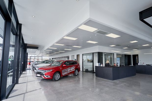 A bright, spacious automotive showroom and reception area with several vehicles displayed indoors. The space features polished grey concrete flooring, extensive black-framed glazing along one side, white ceilings with recessed downlights and suspended rectangular light panels, and a dark reception counter with glazed office partitions.