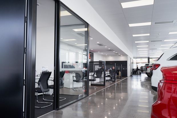 A bright modern automotive showroom and office interior, viewed down a polished showroom corridor. Floor-to-ceiling black-framed glass partitions enclose offices and workstations on the left, while vehicles are displayed along the right side. The space has a suspended white acoustic-tile ceiling with evenly spaced rectangular fluorescent or LED light panels, glossy polished concrete or terrazzo-style flooring, and a clean commercial-industrial appearance.