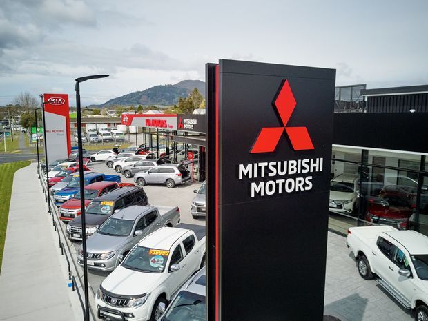 Exterior commercial photograph of a Mitsubishi Motors automotive dealership, dominated by a tall black and red illuminated pylon sign in the foreground. The dealership building and glazed showroom are visible behind it, along with rows of parked vehicles, access paths, street lighting, and a mountainous backdrop. The image is a branded automotive retail setting rather than a residential space.
