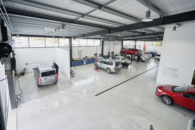 A large, bright automotive workshop or service garage with multiple vehicles, repair bays, workbenches, tool storage, vehicle lifts, and high exposed steel roof framing. The polished light concrete floor and white partition walls create a clean, functional industrial workspace.