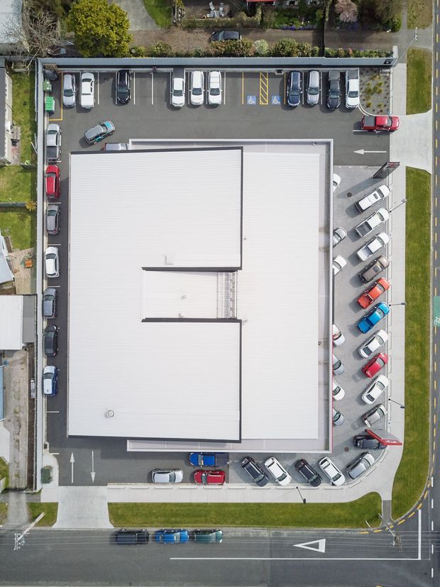 Aerial top-down view of a large commercial building with a bright ribbed metal roof, surrounded by marked surface parking, accessible parking bays, drive lanes, sidewalks, landscaped verges, and adjoining streets. The image focuses on the building roof and its urban commercial site.