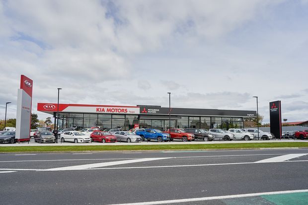 Wide exterior photograph of a low-rise automotive dealership complex viewed from across a multilane road. The building has extensive glazed showroom frontage, a long dark fascia, prominent Kia Motors branding and a separate Mitsubishi pylon sign. Numerous parked vehicles line the forecourt, with cloudy sky and road markings in the foreground.