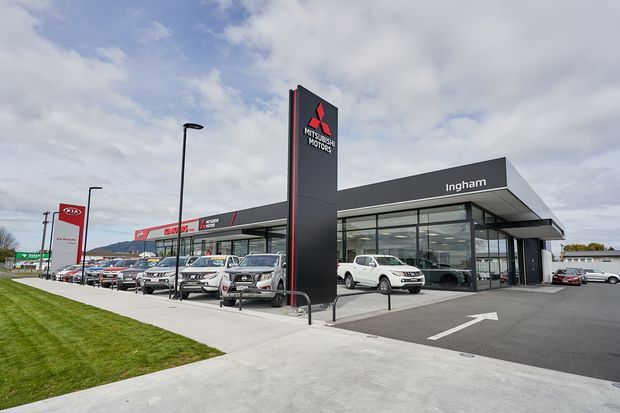 Exterior view of a modern automotive dealership, with a dark grey and glass showroom building, prominent Mitsubishi Motors pylon signage, Ingham branding, vehicle display areas, paved forecourt, and road-access driveway.