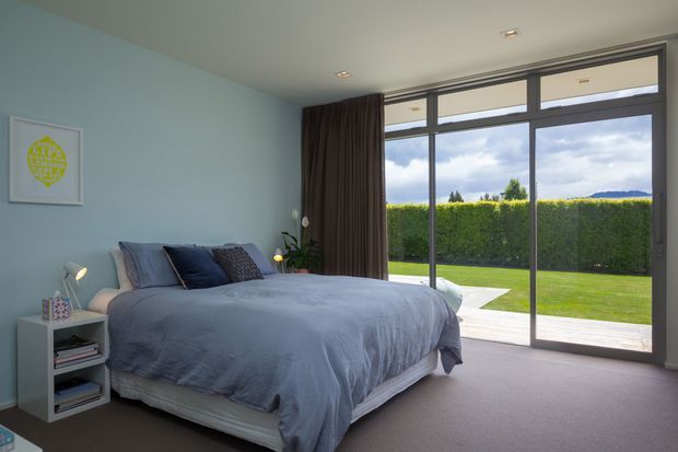 A bright, contemporary bedroom with a large upholstered bed dressed in muted blue-grey bedding, blue and patterned cushions, and a small white bedside table. Pale blue walls, carpeted flooring, dark curtains, and full-height dark-framed glazing create a calm connection to a neatly landscaped garden and distant hills outside.