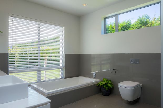 Bright contemporary bathroom with a freestanding-style rectangular bathtub, wall-mounted toilet, large white vanity, grey tiled lower walls, white upper walls, and extensive horizontal window glazing with white blinds. A small indoor plant adds greenery, while the clean-lined fixtures and restrained palette create a modern, airy feel.