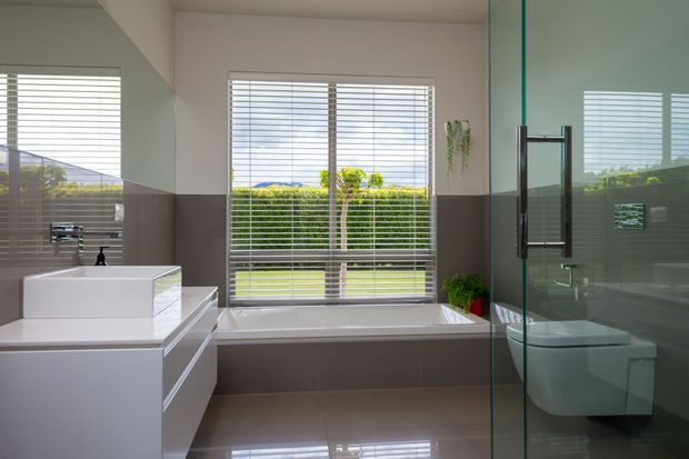 Bright contemporary bathroom with a freestanding bathtub beneath a large window, a floating white vanity, wall-mounted toilet, and frameless glass shower enclosure. Neutral taupe and white finishes, horizontal blinds, and greenery visible outside create a clean, light-filled space.