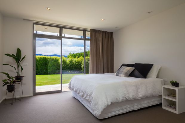 A bright, contemporary bedroom with a large aluminium-framed sliding glass door opening to a neatly landscaped garden. The room features a low-profile upholstered bed with white bedding, neutral layered cushions, full-height taupe curtains, recessed ceiling lights, soft carpet flooring, and a small white bedside unit. Indoor plants add a natural accent.