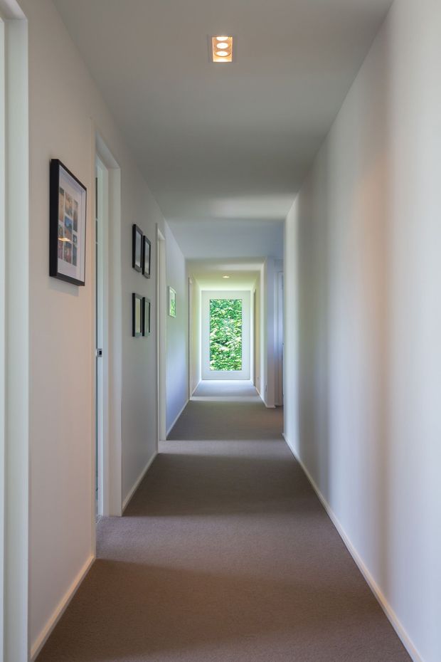 A long, narrow residential hallway with white painted walls and ceiling, grey-beige carpet, white door frames, framed artwork, and recessed ceiling spotlights. Natural light enters through a glazed opening at the far end, with greenery visible beyond.