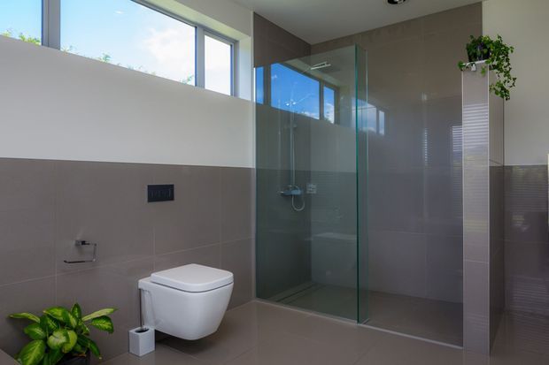 Bright contemporary bathroom with a wall-hung toilet, large walk-in glass shower enclosure, taupe-grey tiled walls and floor, white upper walls, and high horizontal windows. A narrow tiled partition with a trailing plant adds a natural accent.
