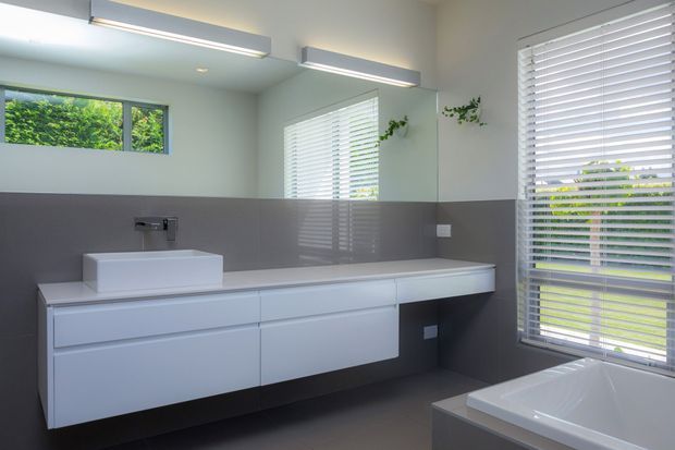 Contemporary bathroom with a long floating white vanity, integrated countertop, above-counter rectangular basin, wall-mounted tapware, large mirror, grey tiled wall finish, and a freestanding white bathtub. Horizontal blinds and greenery are visible through the windows, while linear ceiling-mounted lighting provides even illumination.