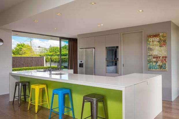 Bright contemporary kitchen with a large white island, vivid lime-green feature panel, and colourful bar stools. The kitchen includes integrated cabinetry, stainless-steel appliances, recessed ceiling lights, and broad glazing overlooking a landscaped outdoor area.