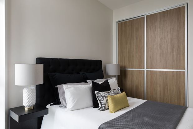A contemporary bedroom with a black tufted upholstered bedhead, white bedding and layered decorative cushions. A black bedside table and white textured lamp sit beside the bed, while full-height sliding wardrobe doors feature warm wood-grain panels with white framing. The room has neutral painted walls and a refined, softly lit appearance.