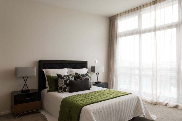 A softly lit contemporary bedroom with a dark upholstered bedhead, layered white and patterned cushions, an olive-green quilted runner, and matching dark bedside furniture. Floor-to-ceiling windows with sheer curtains fill the room with natural light, while neutral walls and carpeting create a calm, refined atmosphere.