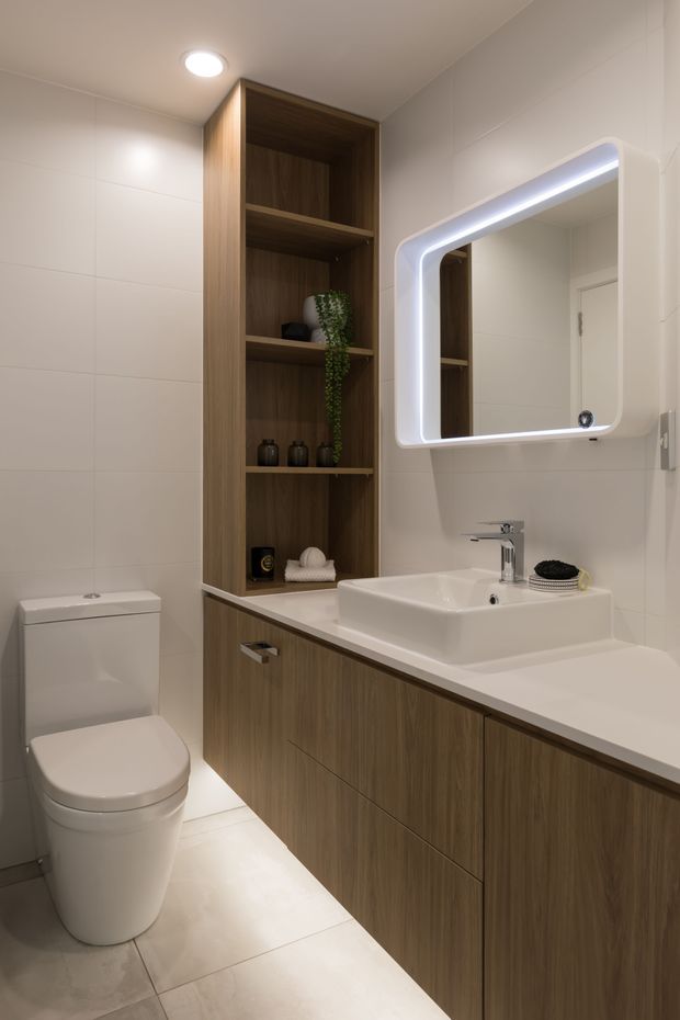 A compact contemporary bathroom with a white toilet, wall-to-wall pale tiles, a timber-look floating vanity with integrated shelving, white basin, chrome tapware, and a rounded-rectangle illuminated mirror. Warm recessed lighting and dark wood joinery create a clean, refined finish.