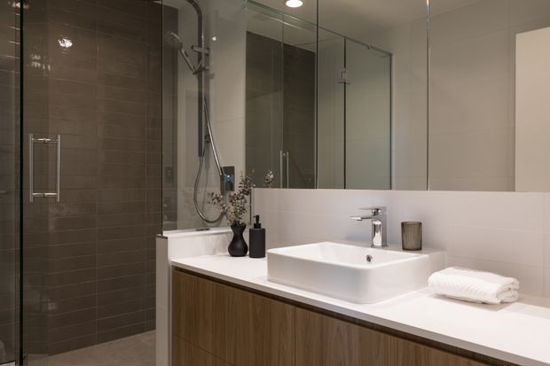 Contemporary bathroom featuring a glass-enclosed shower with dark grey tiled walls, a freestanding white bathtub, and a wall-to-wall timber-look vanity with a white countertop and vessel basin. Large mirrors, chrome tapware, and recessed ceiling lighting create a clean, refined look.
