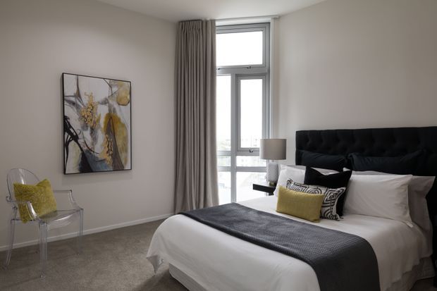 A contemporary bedroom with a neatly made bed featuring a tall black upholstered headboard, white bedding, and a charcoal throw. Floor-to-ceiling grey curtains frame a tall window, while a transparent accent chair with a yellow cushion and abstract artwork add colour. The room has neutral walls, carpeted flooring, and soft natural light.
