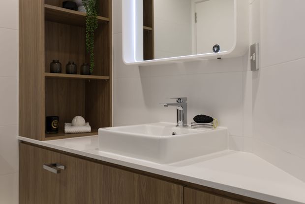 A contemporary bathroom vanity with a white rectangular vessel basin, chrome tapware, integrated white benchtop, illuminated rounded-corner mirror, and warm wood-grain built-in cabinetry with open shelving. White tiled walls and restrained decorative objects create a clean, refined look.