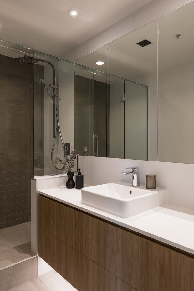 Contemporary bathroom featuring a long floating timber-look vanity with a white solid-surface benchtop, integrated rectangular basin, wall-mounted chrome tapware, large mirrored wall panels, and a frameless glass shower enclosure. The shower has dark grey tiled walls and matching grey floor tiles, with recessed ceiling lighting and minimalist accessories.