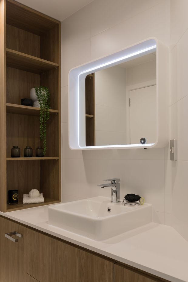 A compact, contemporary bathroom vanity featuring a white rectangular vessel basin, chrome tapware, an illuminated rounded-corner mirror, pale tiled walls, and integrated dark timber-look cabinetry with open shelving and decorative storage.