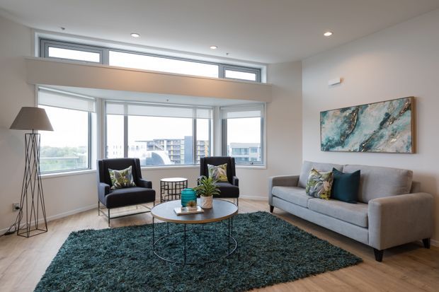 Bright contemporary living room with expansive corner glazing and high clerestory windows, furnished with a grey sofa, two dark lounge chairs, round coffee table, floor lamp, and deep teal textured rug. Light timber flooring, white walls, and abstract artwork create an airy, refined interior with city views beyond the windows.