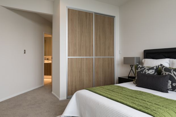 A contemporary bedroom with a neutral palette, carpeted floor, white walls, a dark upholstered bed, patterned accent pillows, and a green quilt. Built-in sliding wardrobe doors feature a light wood-grain finish with white framing. An open doorway leads to a warmly lit bathroom or vanity area.