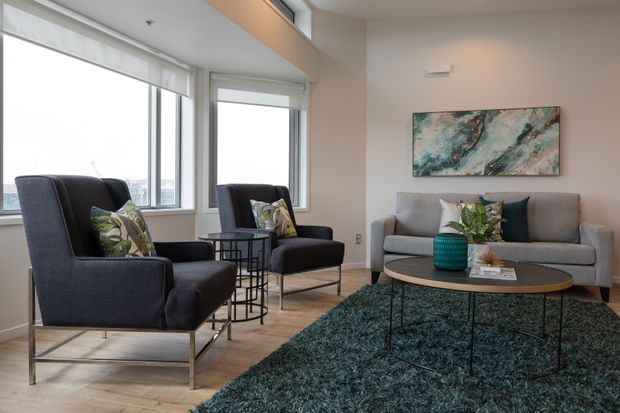 Bright contemporary living room with floor-to-ceiling corner windows, two dark upholstered armchairs, a grey sofa, round coffee table, and deep teal textured rug. Neutral walls and timber-look flooring are complemented by botanical cushions, a teal decorative vase, and abstract artwork.