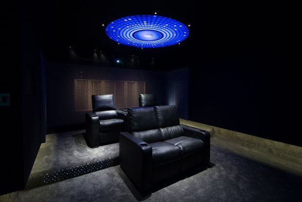 A dark, luxurious private cinema room with two rows of black leather recliner seating on a charcoal carpet. The room has dark walls, a textured wood acoustic feature wall, low-level floor lighting, and a striking circular blue illuminated ceiling feature.