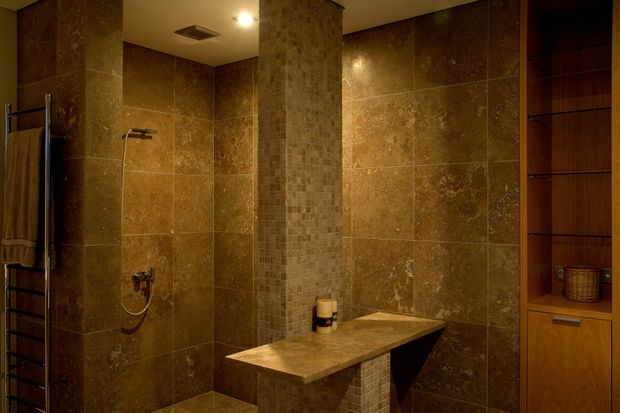 A warm, dimly lit contemporary bathroom clad in large brown stone-look tiles, with a walk-in shower, wall-mounted shower fittings, heated towel rail, tiled feature column, built-in stone bench, and dark timber storage cabinetry.