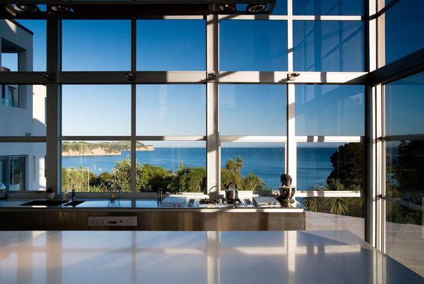 A contemporary kitchen with a long island and stainless-steel appliances, framed by expansive floor-to-ceiling glazing overlooking a blue coastal bay. The space has dark structural framing, reflective pale surfaces, and abundant natural light.