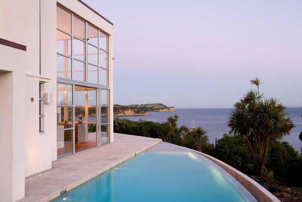 A contemporary coastal home exterior with a large floor-to-ceiling glazed facade opening onto a pale stone terrace and an infinity-edge swimming pool. The pool overlooks a calm ocean bay with a vegetated headland, while palms and coastal planting frame the scene. The image has a refined, airy sunset atmosphere.
