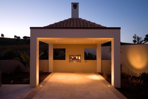 A symmetrical covered outdoor entertaining area at dusk, attached to a stucco building with a terracotta tiled hipped roof and chimney. Square columns frame a tiled patio with recessed lighting, a central built-in fireplace, and landscaped planting along the perimeter walls.