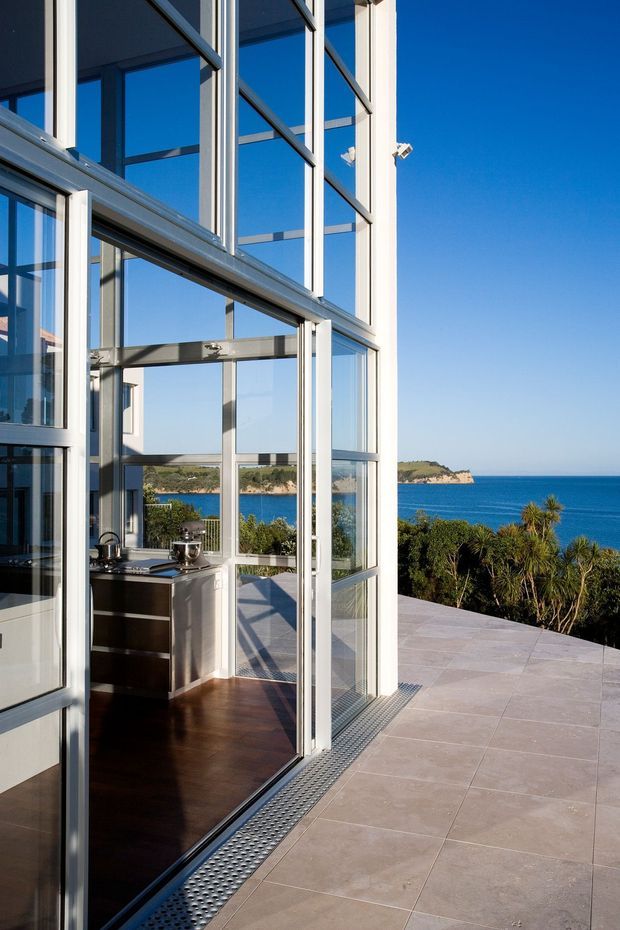 A modern coastal residence with expansive floor-to-ceiling glazed walls and slender light-coloured metal window frames. The view is from a tiled outdoor terrace looking into a compact kitchen through open glass doors, with dark timber flooring and stainless-steel cabinetry or appliances visible inside. Beyond the terrace are landscaped coastal plants, a vivid blue sea, and a rocky headland.