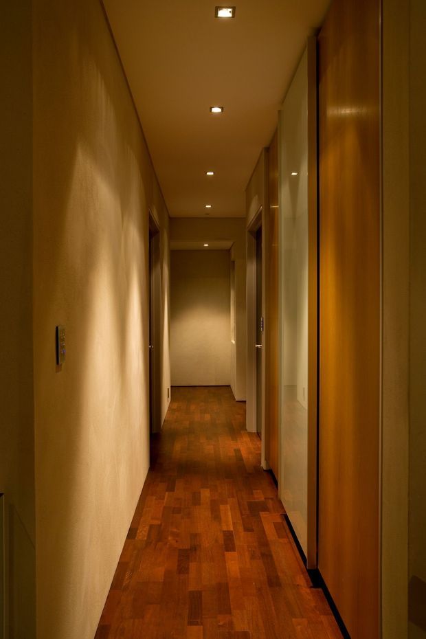 A long, narrow residential hallway with warm recessed ceiling lighting, smooth neutral plaster walls, multiple timber doorways and built-in timber panels, and a richly toned parquet timber floor. The corridor has a quiet, enclosed and softly lit atmosphere.