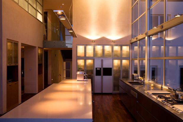 A dramatic, high-ceiling contemporary kitchen with a long pale island, dark built-in cabinetry, stainless-steel appliances, and extensive floor-to-ceiling glazing. Warm under-cabinet and ceiling lighting contrasts with the cool blue exterior light, while a mezzanine-like bridge and tall internal volume create a spacious, multi-level feel.