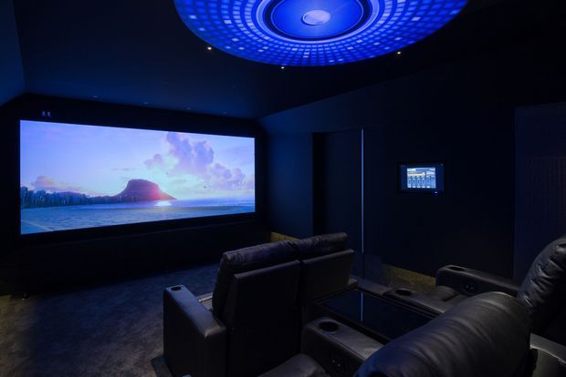 A dark, dedicated home cinema with tiered black leather recliner seating, a large wall-mounted projection screen displaying a landscape scene, a small control panel on the side wall, and a dramatic circular blue illuminated ceiling feature.