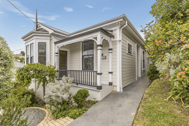 Exterior view of a compact heritage-style single-storey weatherboard house with ornate white trim, a projecting bay window, covered front porch, dark railing, and a narrow path through a landscaped garden. The home has a pitched roof and traditional detailing, photographed in bright daylight.
