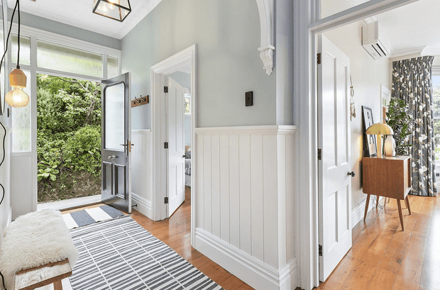 Bright traditional-style entry hall with white-painted wall panelling and decorative trim, warm timber flooring, a glazed exterior door opening to greenery, and a patterned runner. An adjoining furnished room is visible through the doorway on the right, with a timber cabinet, lamp, and patterned curtains.