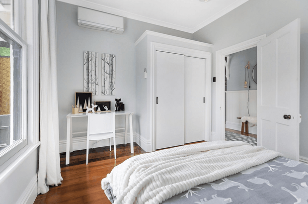 Bright, tidy bedroom with pale grey walls, white trim and doors, warm timber flooring, and a bed in the foreground dressed in a grey patterned cover and chunky white textured throw. A compact white desk and chair sit beside the window, with minimalist artwork and decor above. Built-in sliding wardrobe doors, an open doorway to an adjacent space, and a wall-mounted air conditioner are visible.